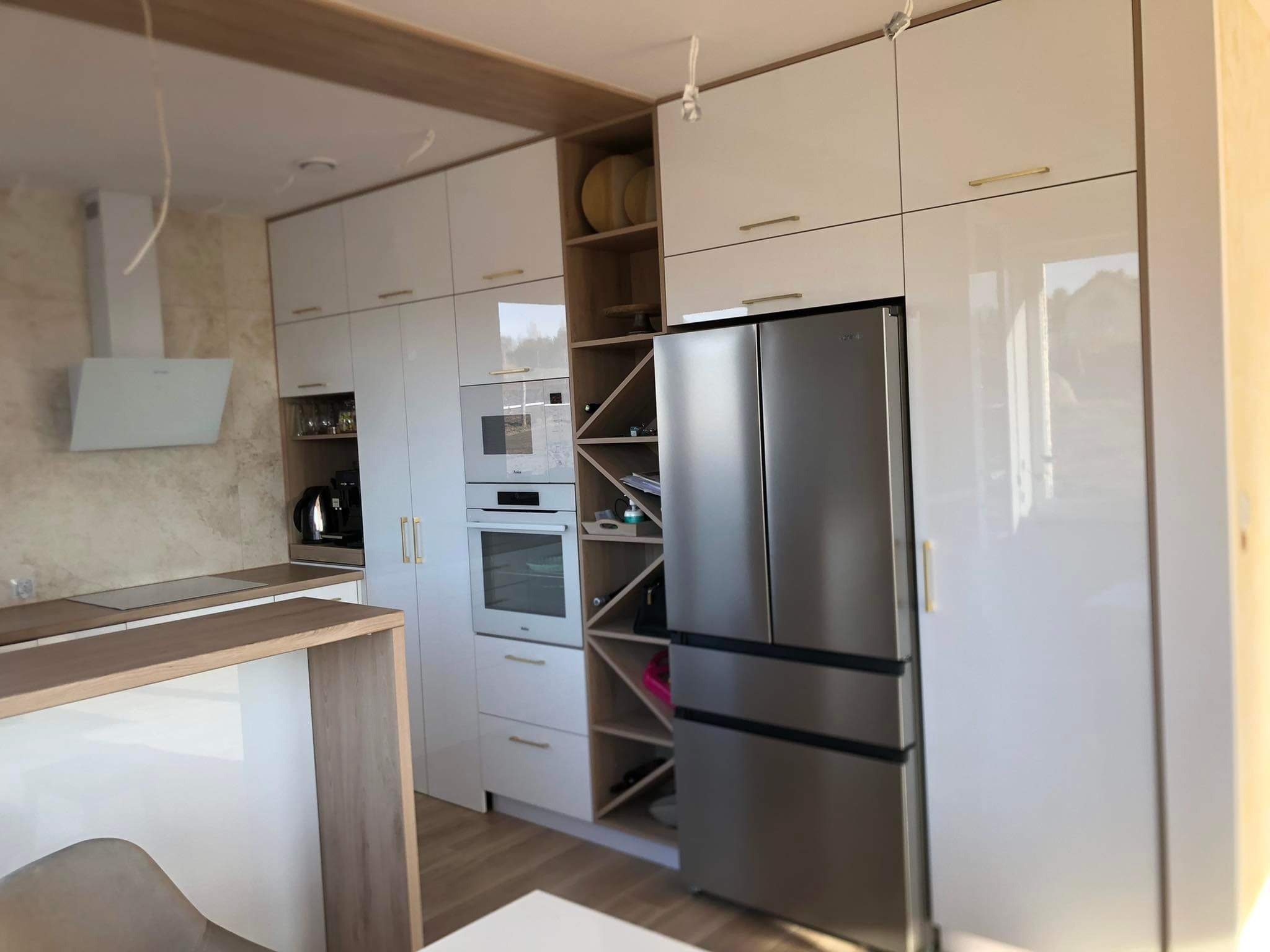 Nowoczesna kuchnia z białymi, lakierowanymi frontami, drewnianymi akcentami i srebrną lodówką side-by-side, widok na wyspę kuchenną.