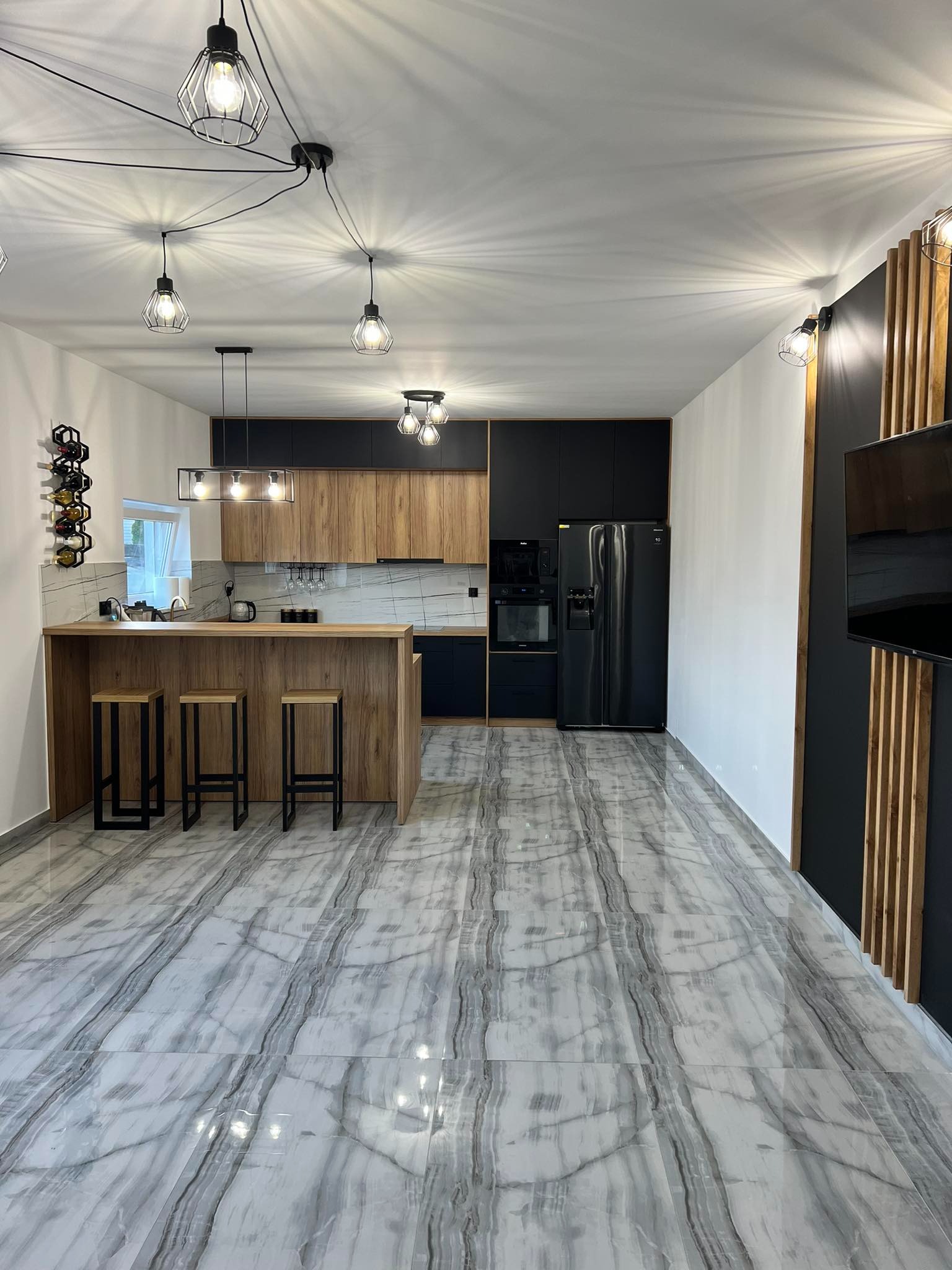 Nowoczesna kuchnia z drewnianymi frontami szafek dolnych i czarnymi górnymi, wyspa z trzema hokerami, marmurowa podłoga i dekoracyjna ściana z drewnianymi listwami.