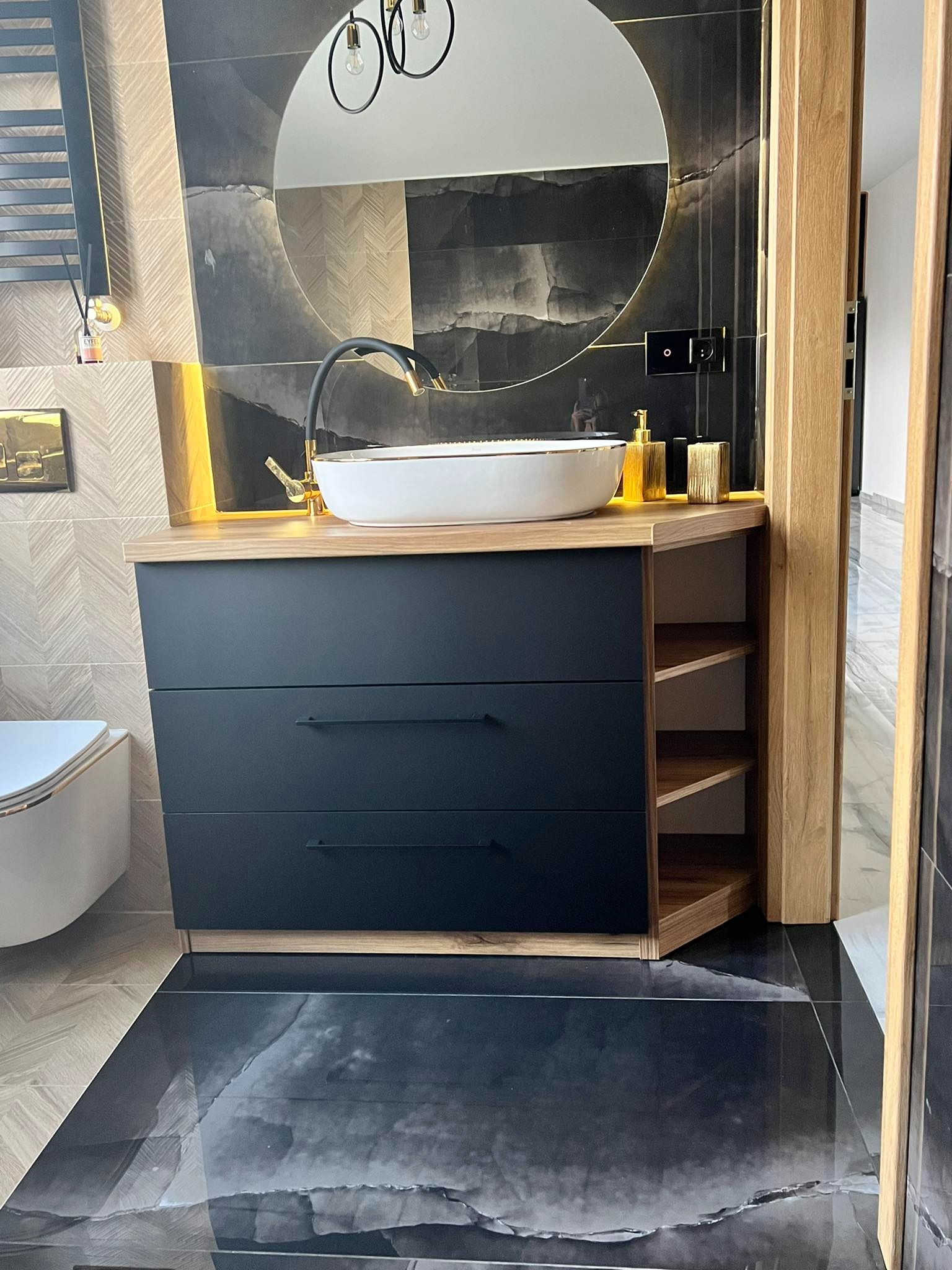 Nowoczesna łazienka z czarną szafką pod umywalkę, drewnianym blatem i półkami, okrągłym lustrem z podświetleniem oraz czarno-białymi płytkami na ścianie.