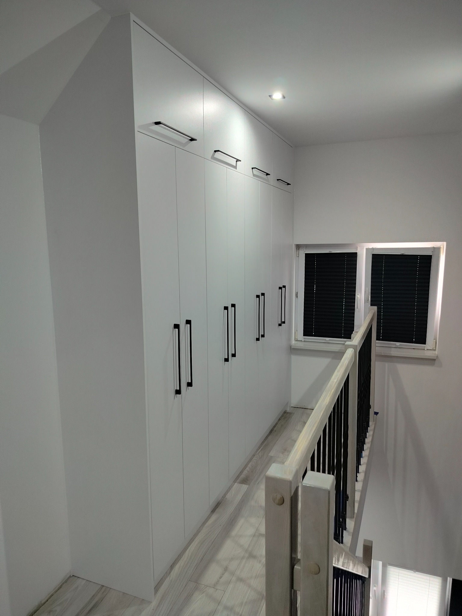 Biała szafa wnękowa z czarnymi uchwytami, zamontowana w korytarzu przy schodach, widoczna z góry, z oknem z żaluzjami w tle.