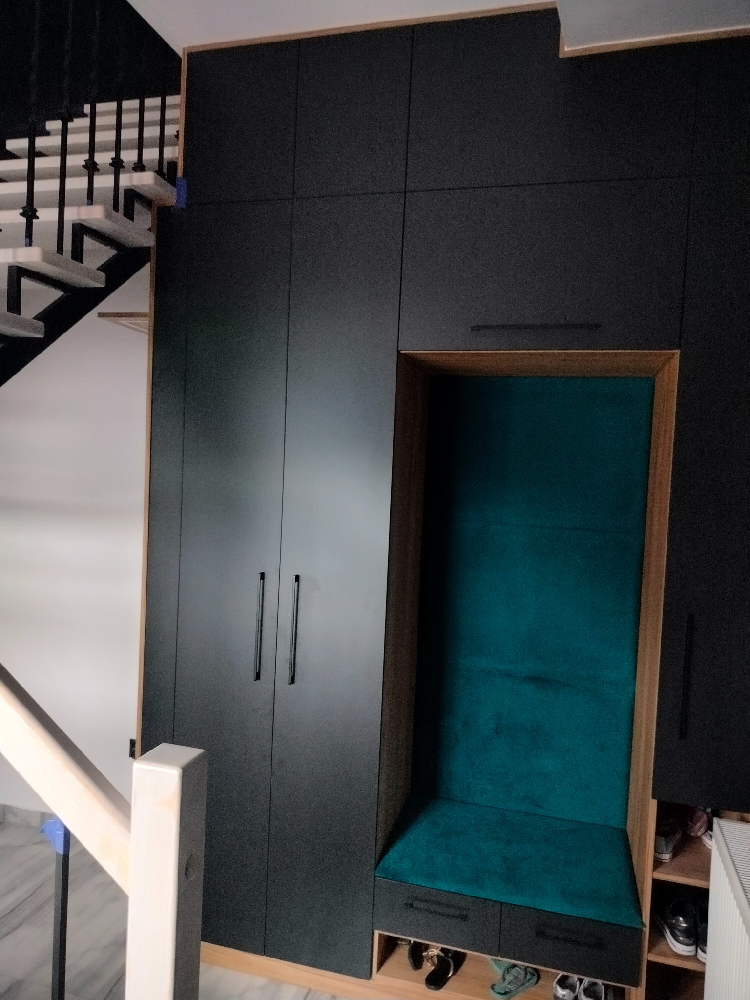 Ciemna zabudowa meblowa z siedziskiem obitym turkusowym welurem, wkomponowana w przestrzeń pod schodami, z widocznymi butami na półkach.