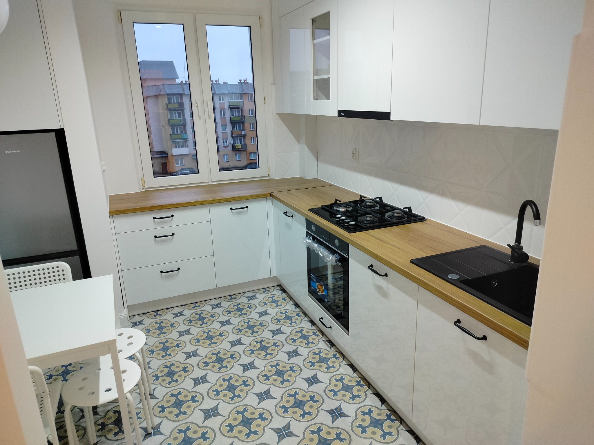 Jasna kuchnia z białymi szafkami, drewnianym blatem i wzorzystą podłogą. Widok na blok z okna.