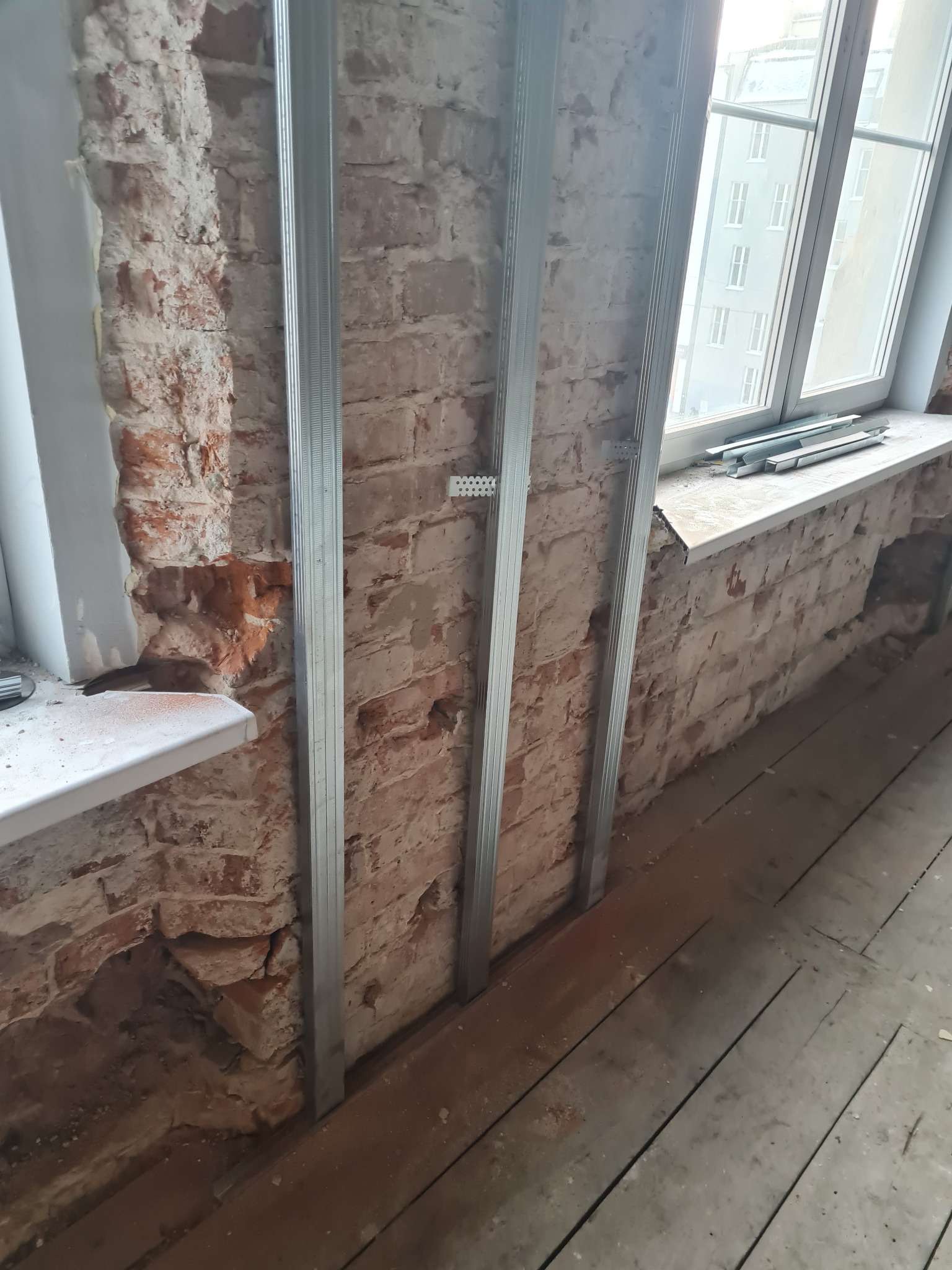 Przygotowanie do montażu płyty karton gips na ścianie z czerwonej cegły, widoczne metalowe profile pionowe przy oknie.