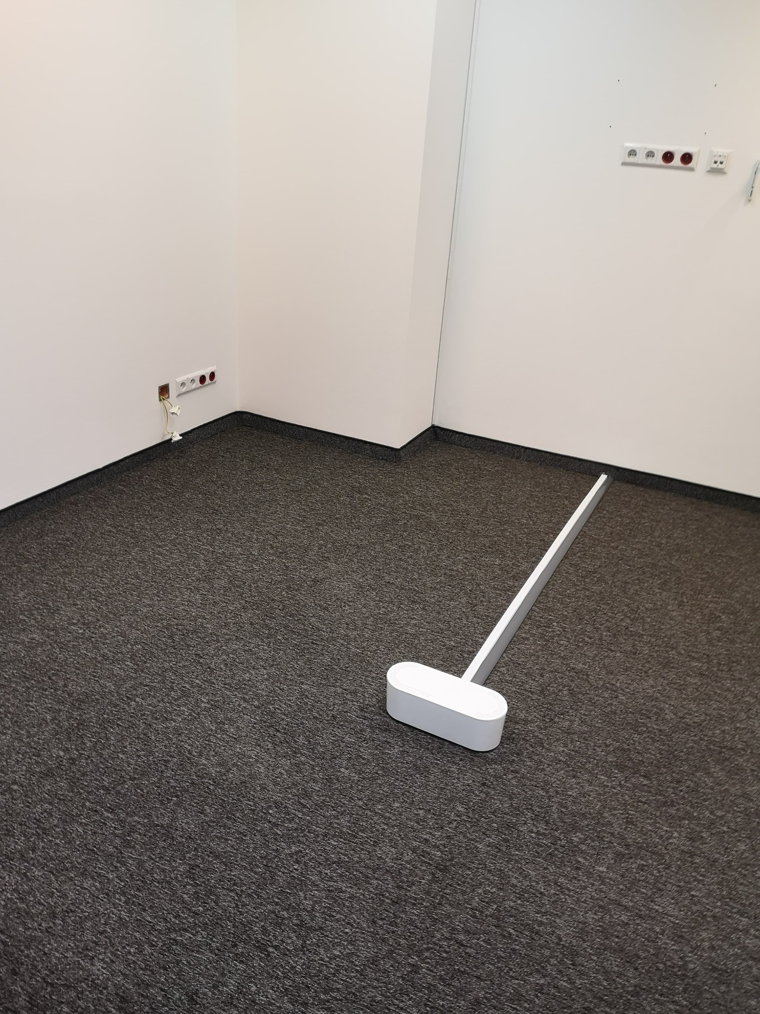 Pusty, pomalowany na biało pokój biurowy z szarą wykładziną dywanową i listwą przypodłogową, widoczne gniazdka elektryczne i włączniki światła na ścianie, na podłodze biały próg