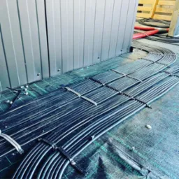 Równolegle ułożone wiązki czarnych kabli elektrycznych, zabezpieczone białymi opaskami, prowadzone po zielonej folii ochronnej wzdłuż szarej metalowej ściany.