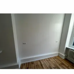 Wnętrze świeżo pomalowanego pokoju z drewnianą podłogą, widoczne gniazdka elektryczne i biały grzejnik pod oknem.