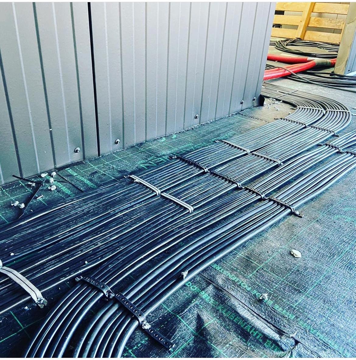 Równolegle ułożone wiązki czarnych kabli elektrycznych, zabezpieczone białymi opaskami, prowadzone po zielonej folii ochronnej wzdłuż szarej metalowej ściany.