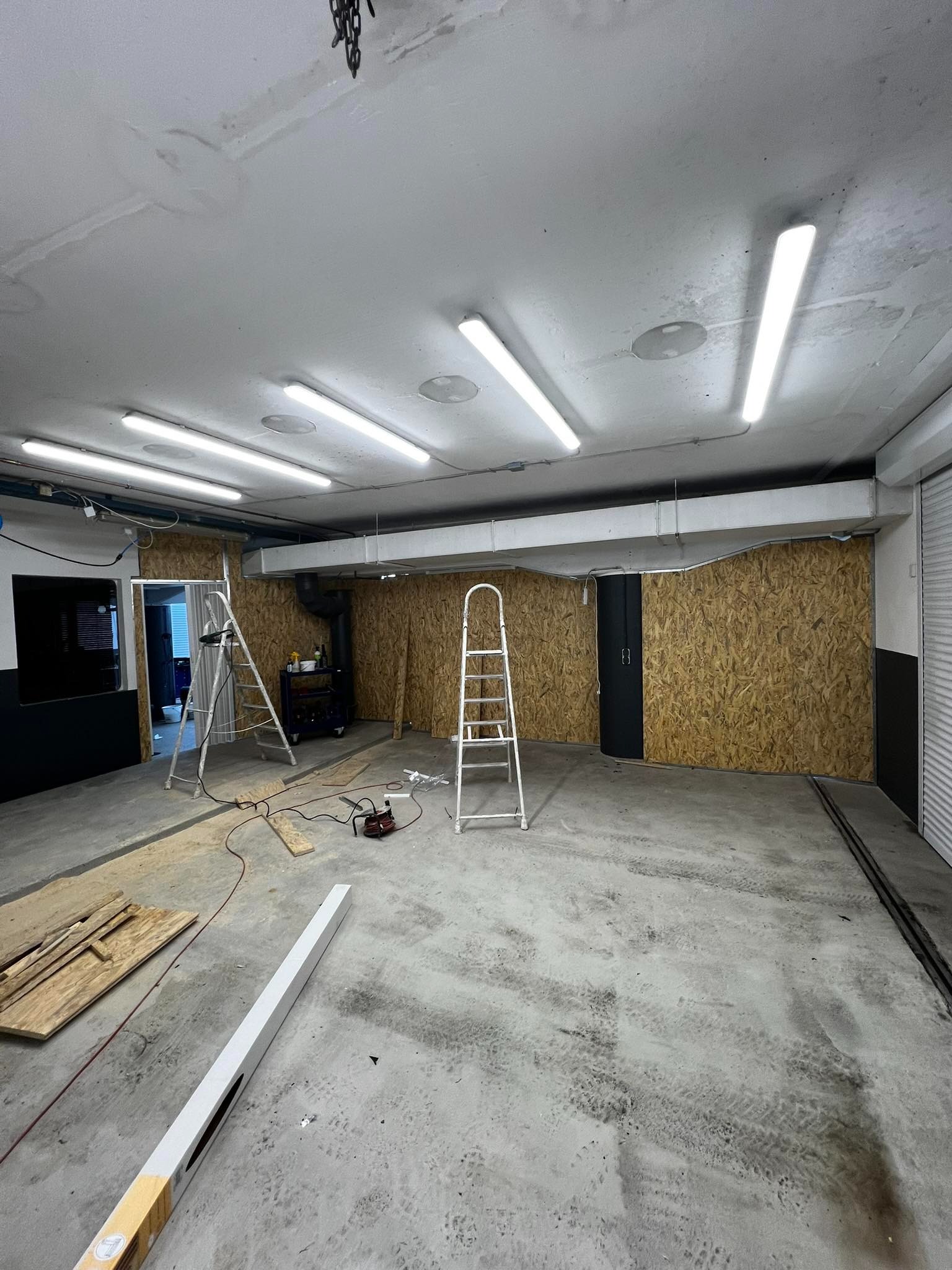 Wnętrze garażu w trakcie adaptacji: ściany wyłożone płytami OSB, dwie drabiny, narzędzia i materiały budowlane na posadzce, widoczne instalacje pod sufitem.