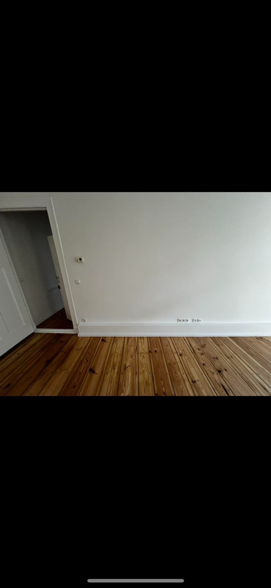 Pusty pokój po odświeżeniu: białe ściany, odnowiona drewniana podłoga, widoczne gniazdka elektryczne przy podłodze, otwarte białe drzwi w tle.