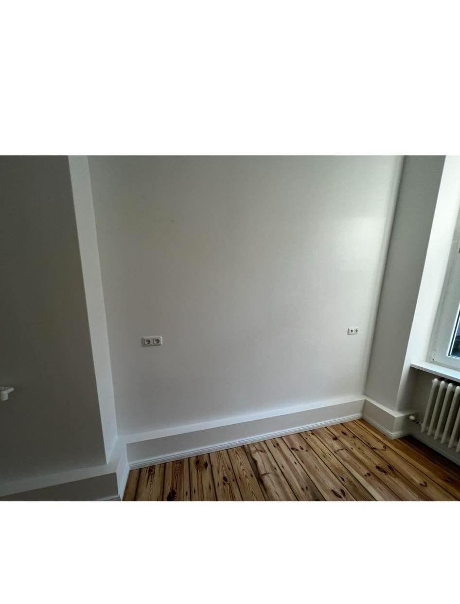 Wnętrze świeżo pomalowanego pokoju z drewnianą podłogą, widoczne gniazdka elektryczne i biały grzejnik pod oknem.
