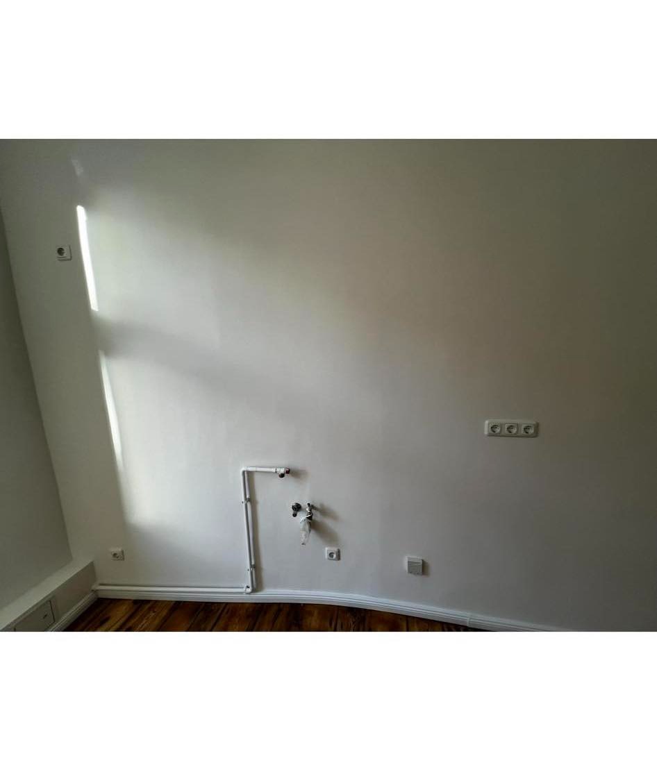 Biała ściana po remoncie z widocznymi gniazdkami elektrycznymi, pionową instalacją wodną z zaworami i drewnianą podłogą w odcieniach brązu.