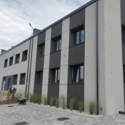 Dwukondygnacyjny budynek biurowy z elewacją w pionowe pasy szarości, z oknami w czarnych ramach, otoczony kostką brukową i niską zielenią, z widocznym w tle kominem fabrycznym.