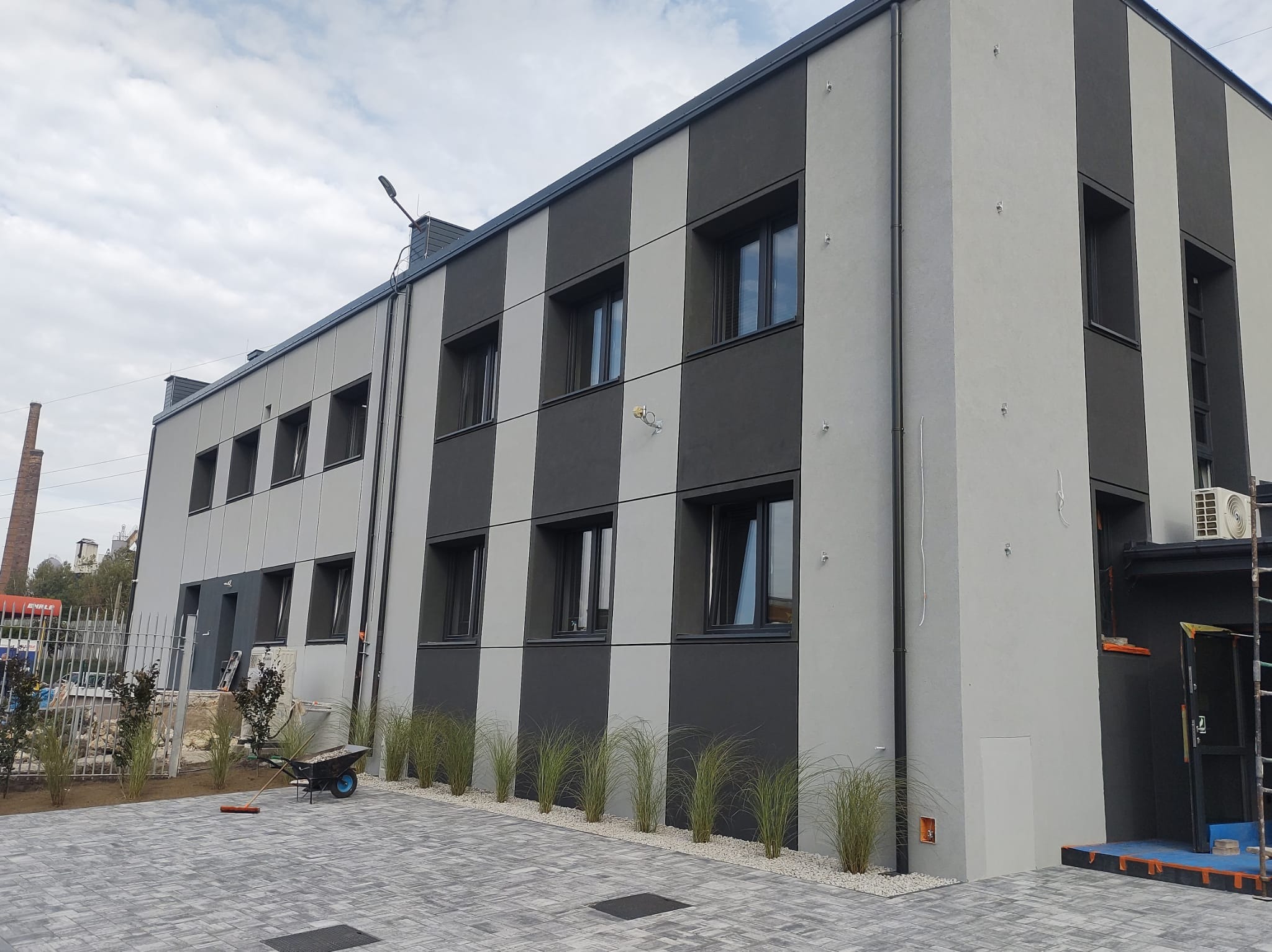 Dwukondygnacyjny budynek biurowy z elewacją w pionowe pasy szarości, z oknami w czarnych ramach, otoczony kostką brukową i niską zielenią, z widocznym w tle kominem fabrycznym.