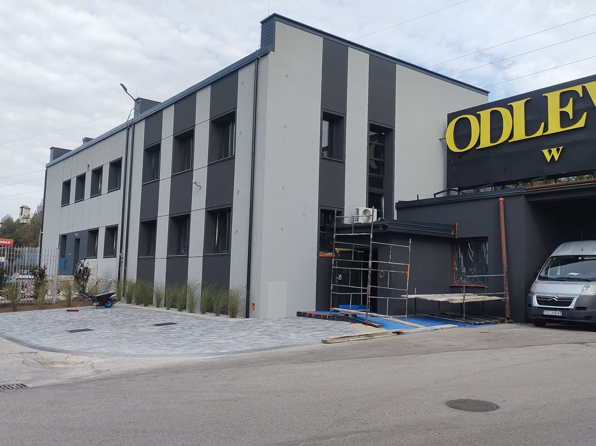 Budynek z elewacją w szaro-białe pasy, częściowo w trakcie prac wykończeniowych, widoczne rusztowania i zaparkowany bus, teren wokół budynku z nową kostką brukową i nasadzeniami.