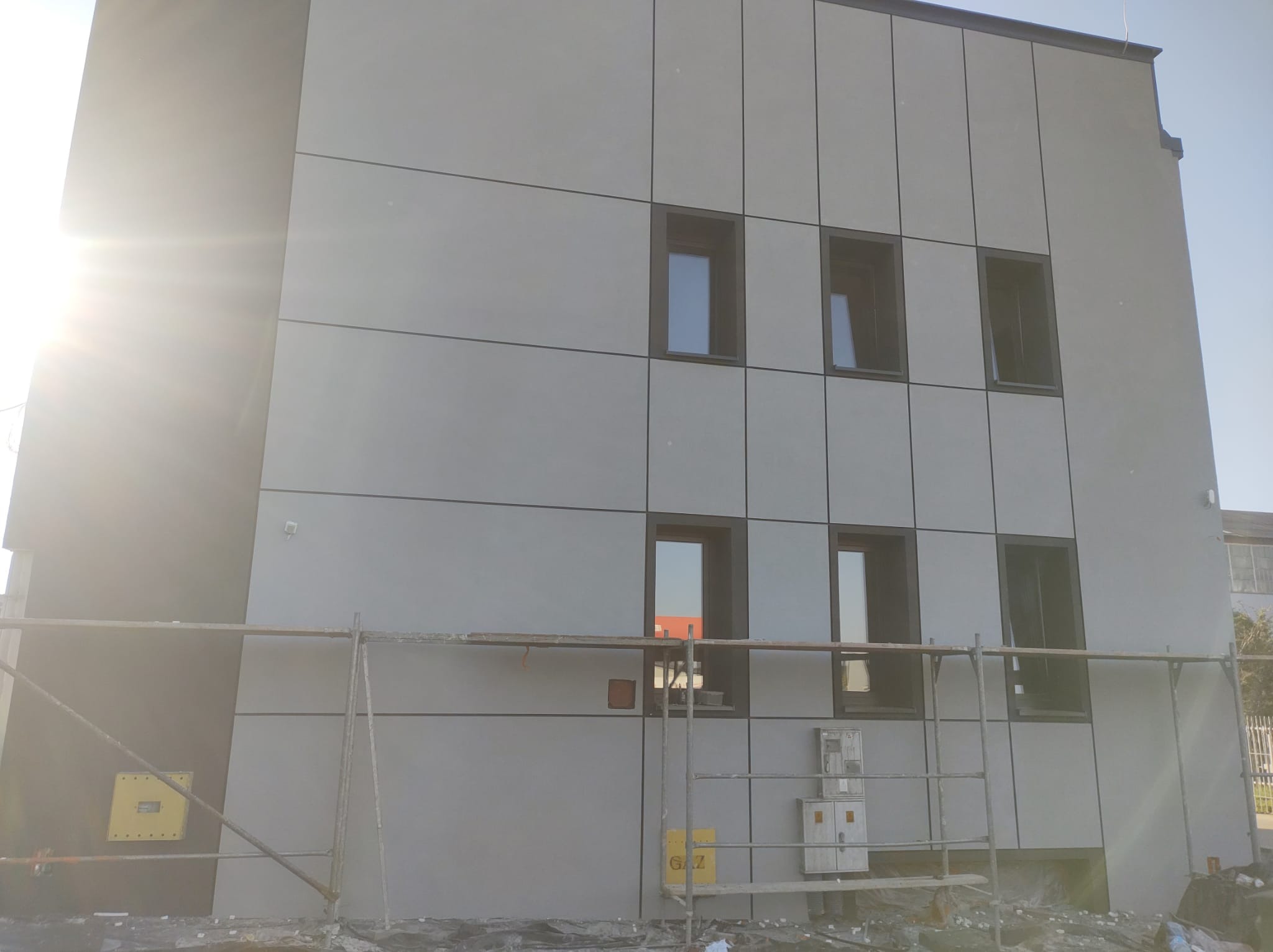 Budynek z szarą elewacją panelową, czarne ramy okienne, widoczne rusztowanie, skrzynka gazowa z napisem GAZ.