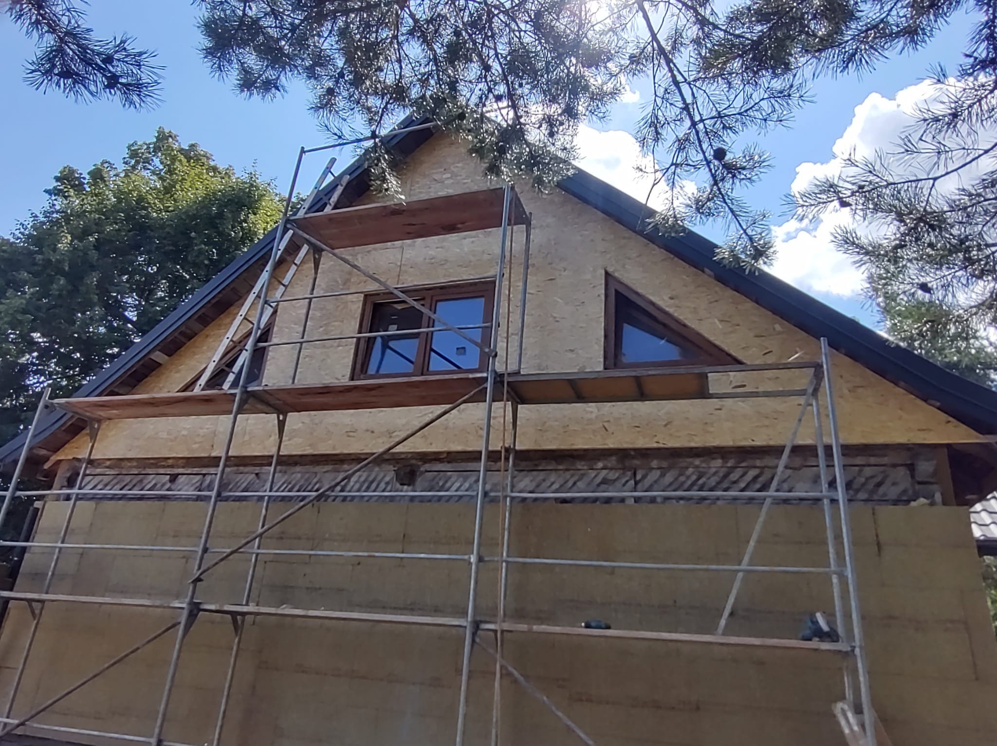 Dom w trakcie ocieplania ścian zewnętrznych, widoczne rusztowania, płyty OSB na szczycie i warstwa izolacyjna na dole ściany, fragmenty drzew na tle błękitnego nieba.