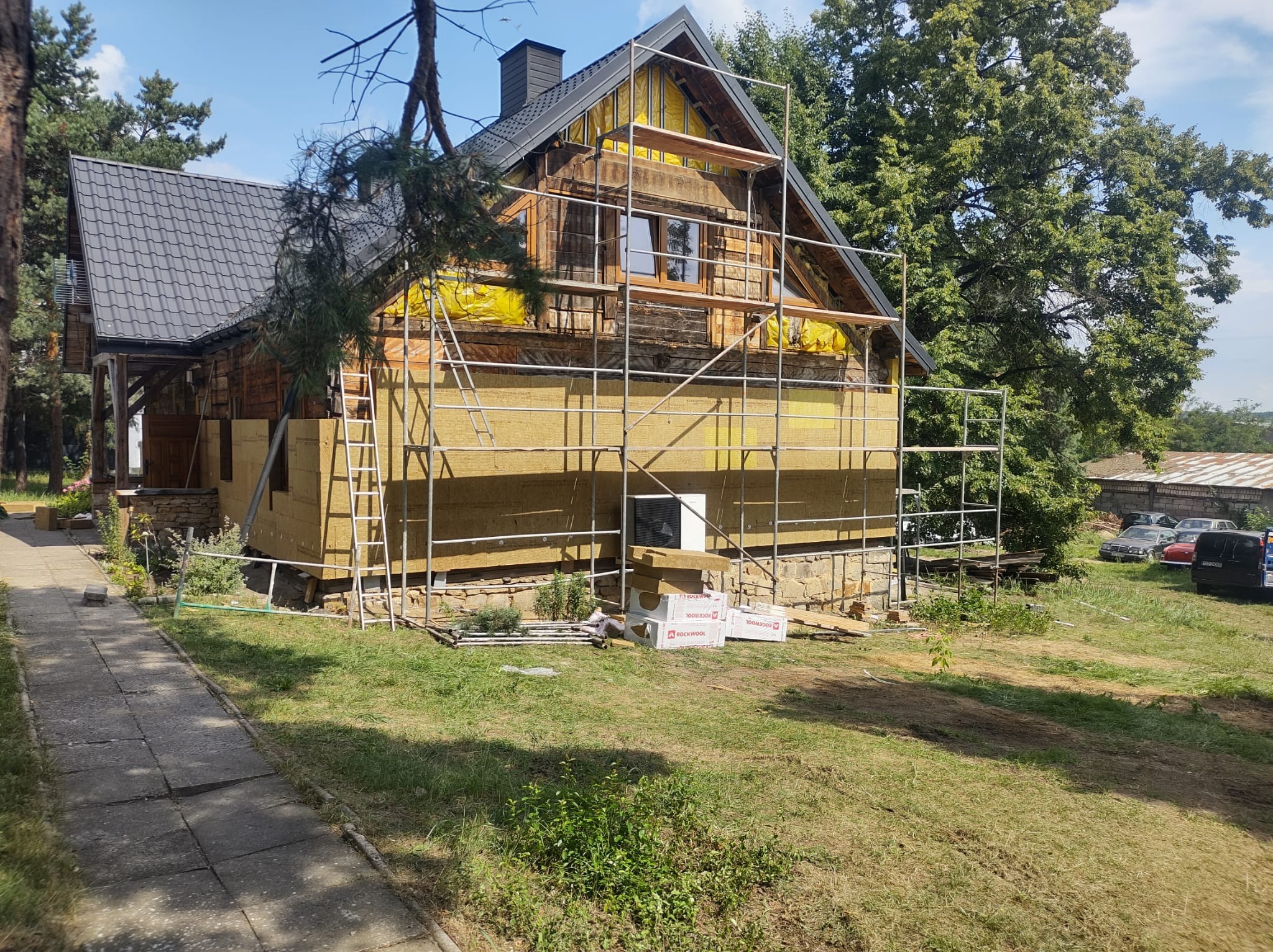 Dom z drewna i kamienia w trakcie ocieplania, widoczne rusztowania, żółta folia i wełna mineralna, paczki wełny Rockwool leżą na trawie.
