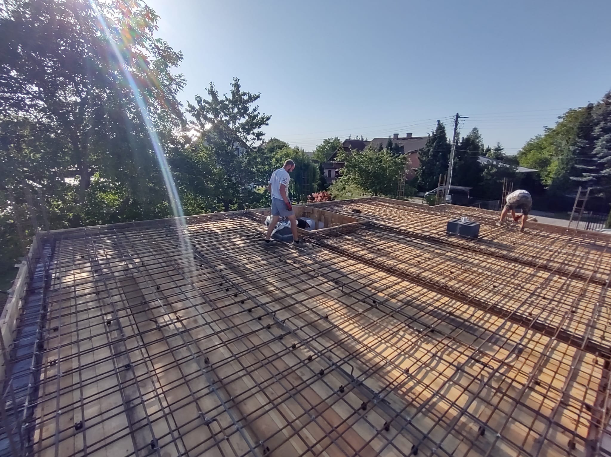 Dwóch pracowników układa siatkę zbrojeniową na drewnianym szalunku stropu, widok z góry, w tle zabudowa jednorodzinna i zieleń drzew, promienie słoneczne.