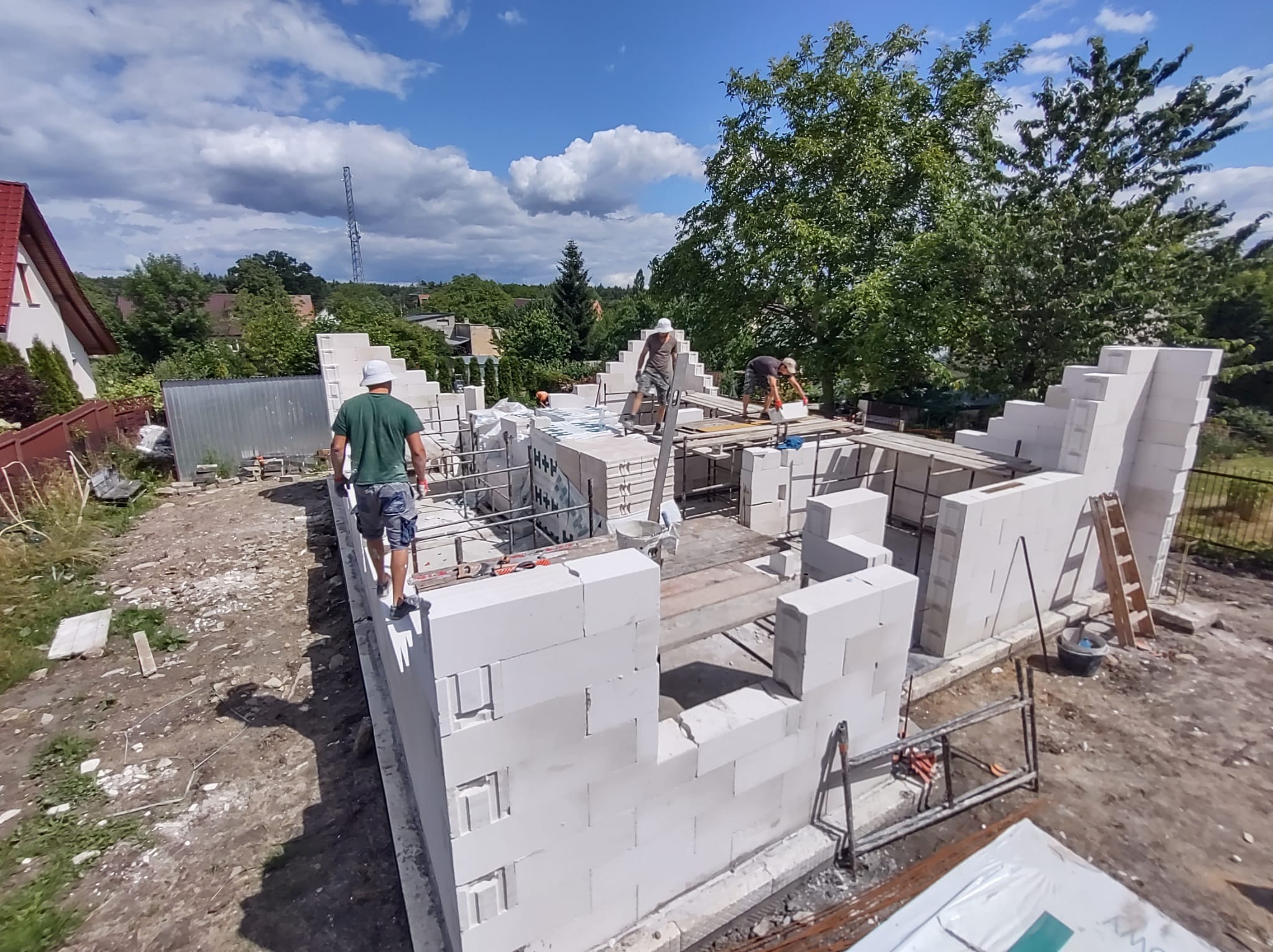 Budowa domu jednorodzinnego z białych bloczków, widoczni robotnicy w kaskach, rusztowania i narzędzia, w tle zieleń drzew i błękitne niebo z chmurami.