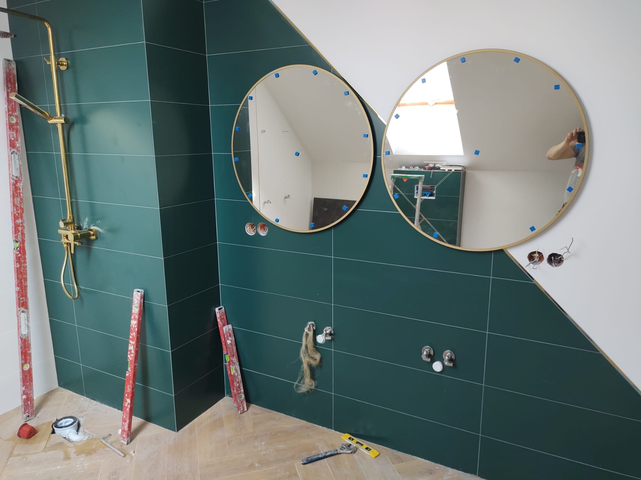 Łazienka w trakcie wykończenia: zielone płytki, złota armatura prysznicowa, dwa okrągłe lustra z niebieskimi podkładkami, instalacja wodna w ścianie, poziomice i narzędzia na podłodze z jasnej...