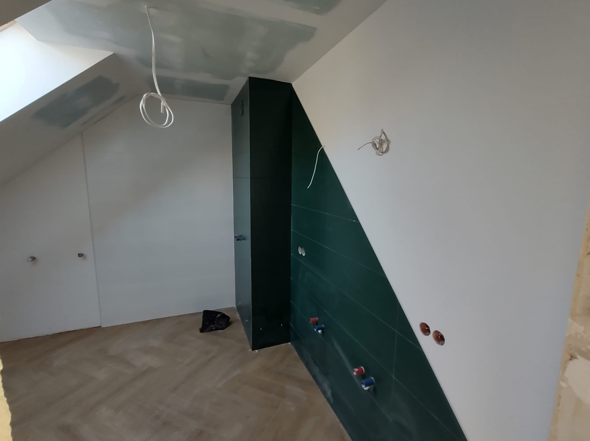 Łazienka na poddaszu w trakcie remontu: zielona ściana z poziomych płytek, sufit i ściany częściowo pomalowane na biało, widoczne instalacje wodne i elektryczne, podłoga wyłożona parkietem...