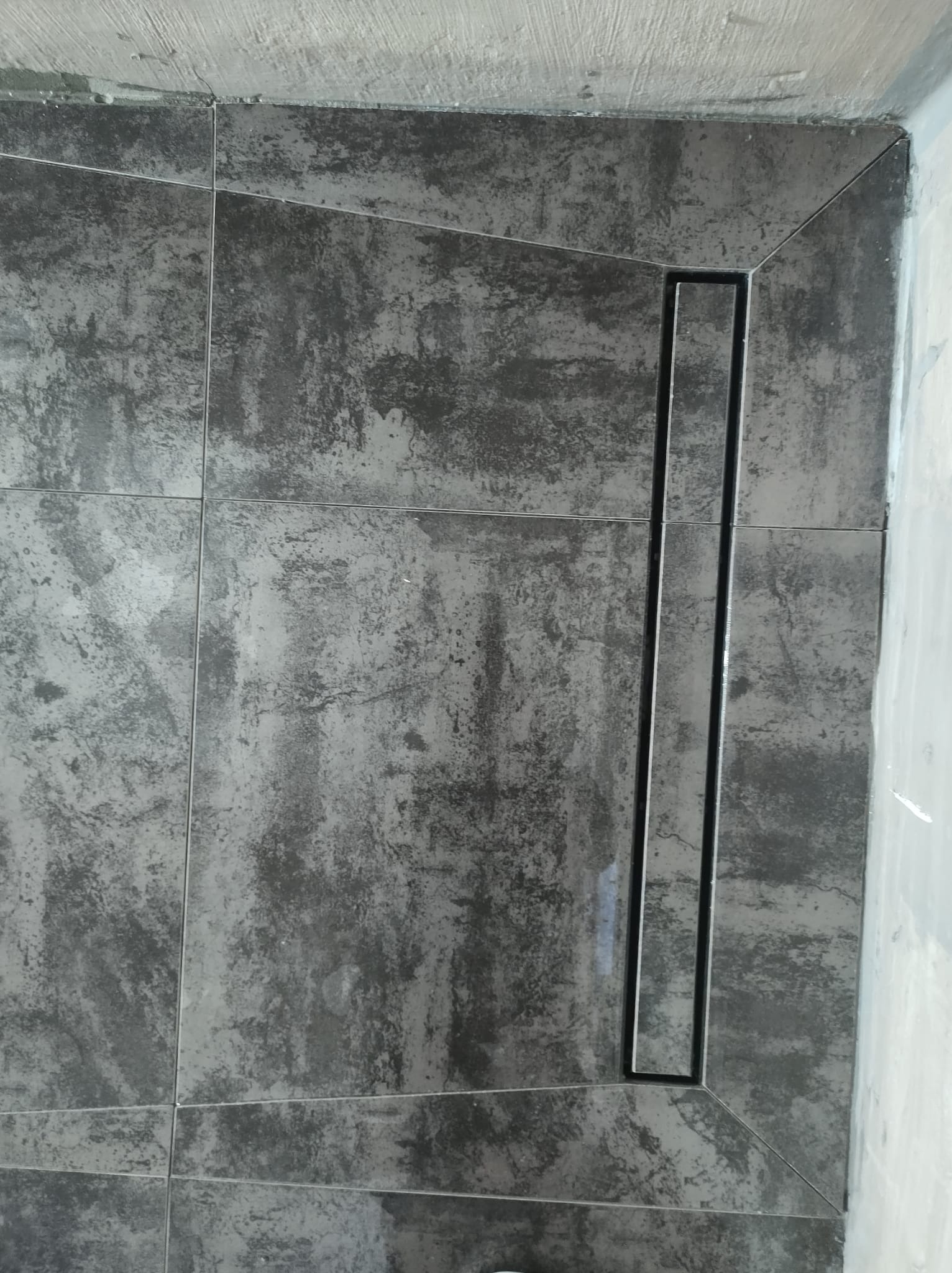 Ciemnoszare płytki imitujące beton, ułożone na podłodze i częściowo na ścianie, z zamontowanym liniowym odpływem prysznicowym z czarną ramką.
