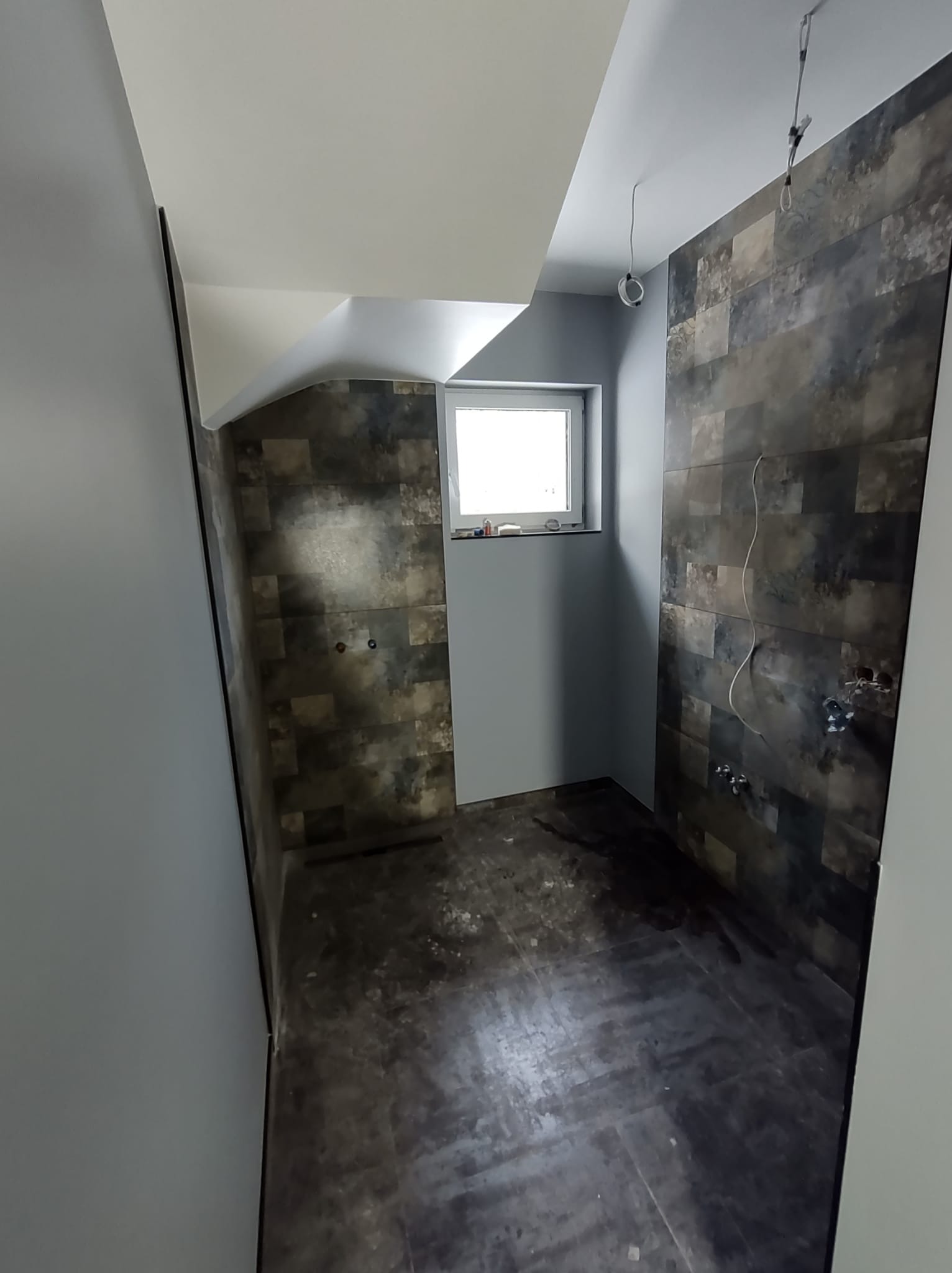 Wnętrze łazienki w trakcie remontu, widoczne ułożone ciemne płytki imitujące kamień na ścianach, surowe instalacje elektryczne i wodne, oraz okno z parapetem. Podłoga częściowo zabezpieczona folią.