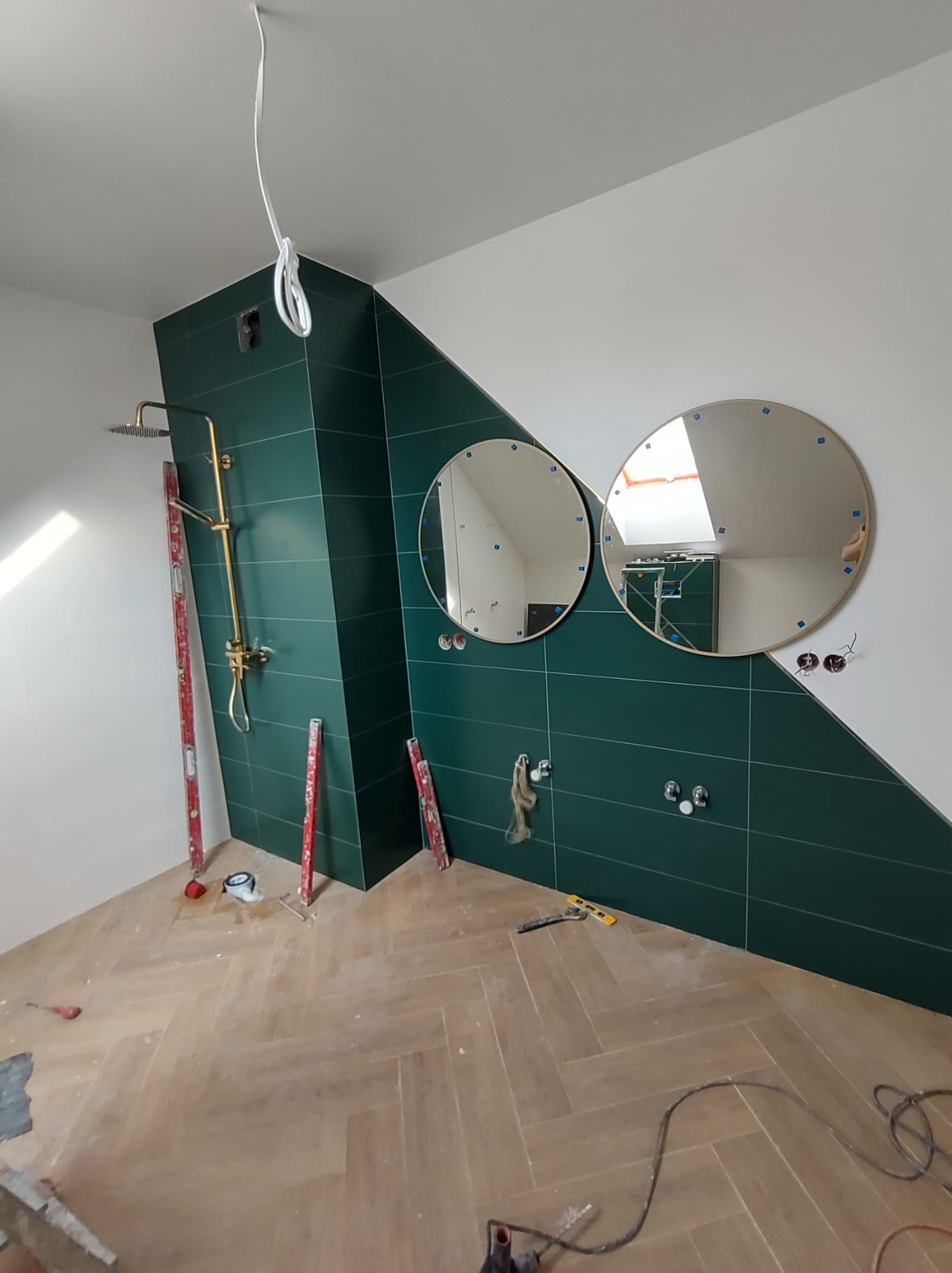 Świeżo wykafelkowana łazienka z zielonymi płytkami, złotą armaturą prysznicową i dwoma okrągłymi lustrami z niebieskimi znacznikami, drewniana podłoga w jodełkę, poziomice i narzędzia na podłodze...