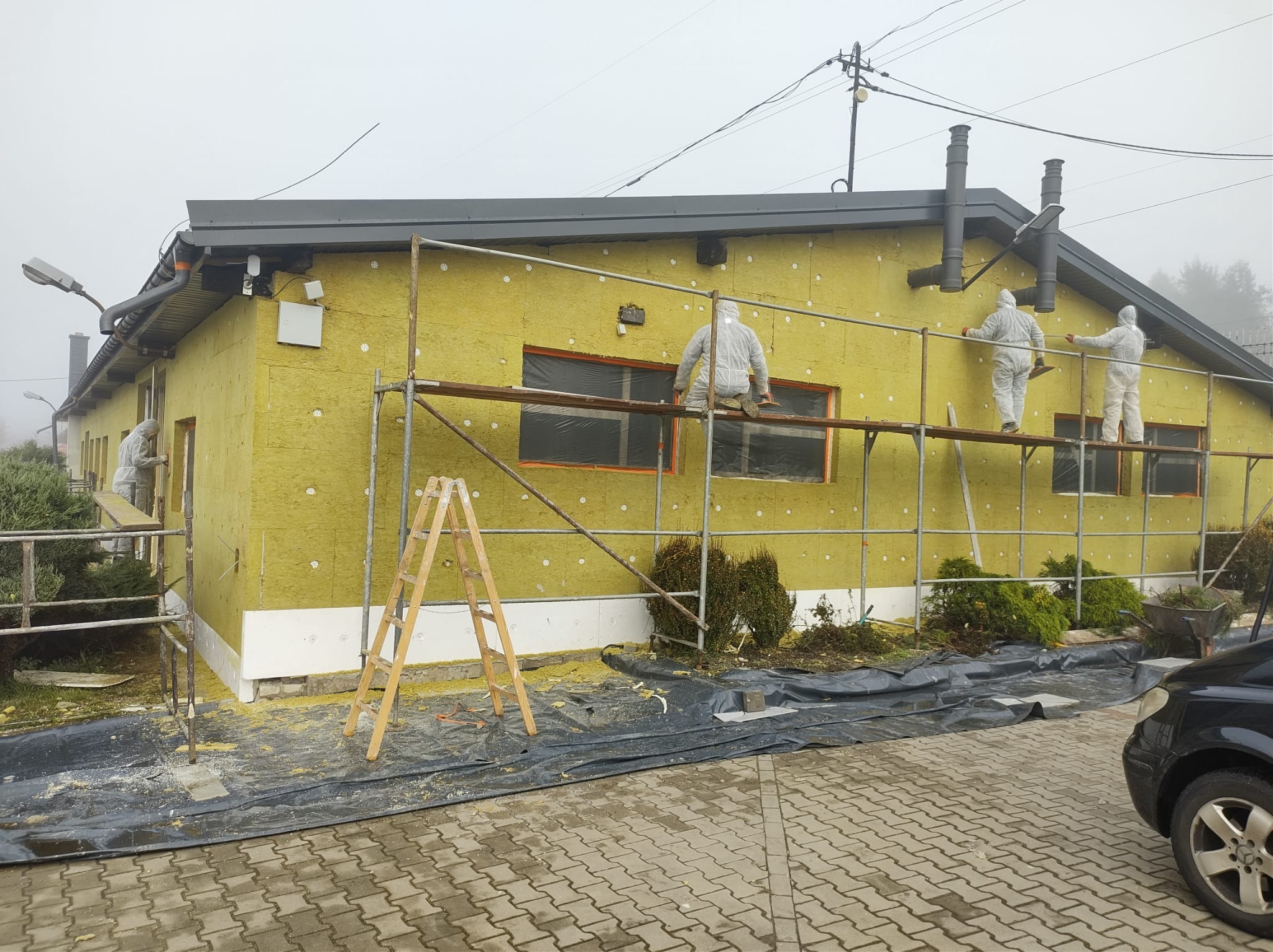 Prace ociepleniowe budynku z żółtą wełną mineralną, pracownicy w białych kombinezonach na rusztowaniu, widoczne okna zabezpieczone folią oraz drewniana drabina