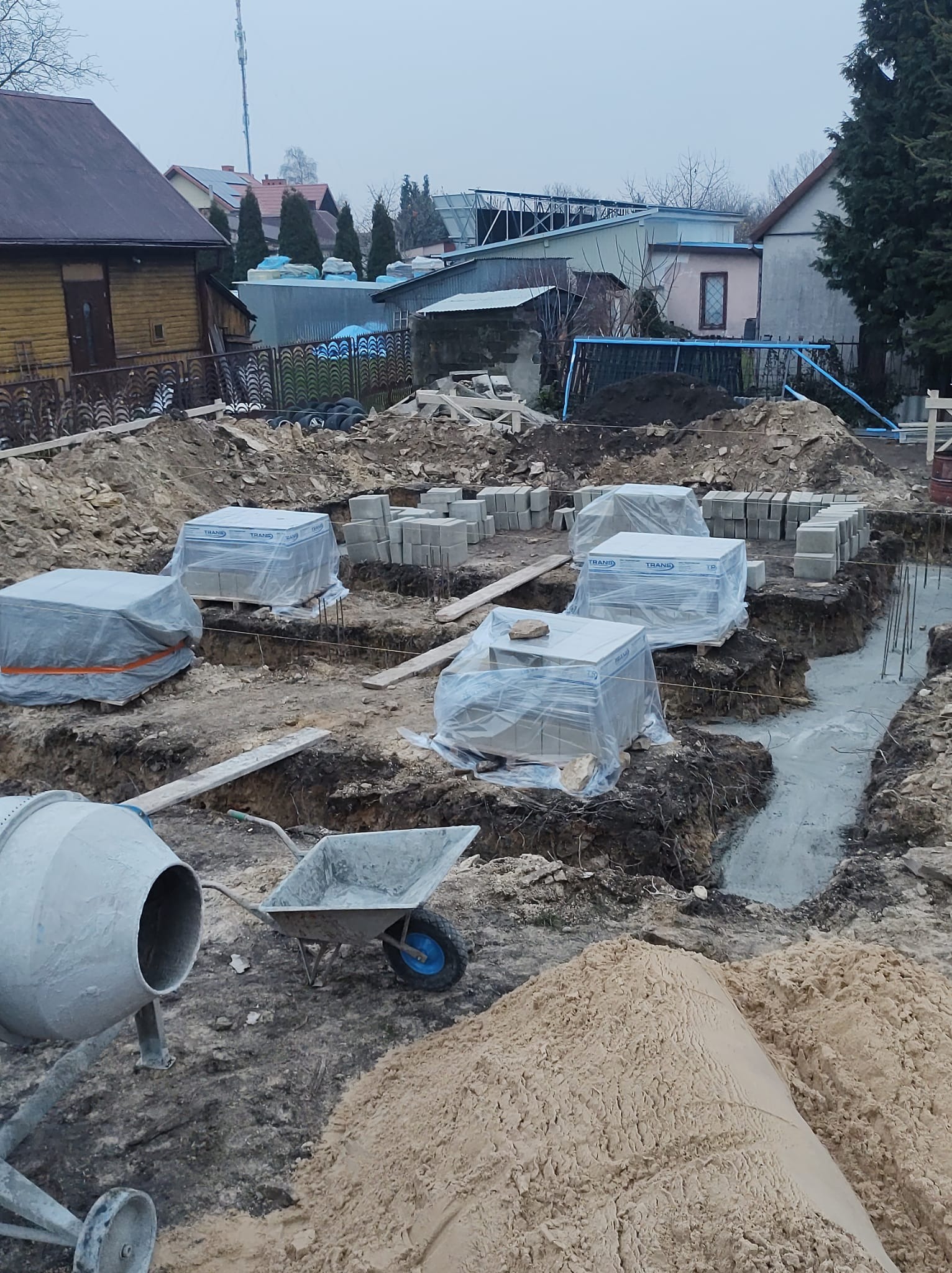 Przygotowanie fundamentów pod budowę domu: wykopane rowy zalane betonem, palety z bloczkami betonowymi zabezpieczone folią, betoniarka i taczka w tle, widok na działkę z domami.