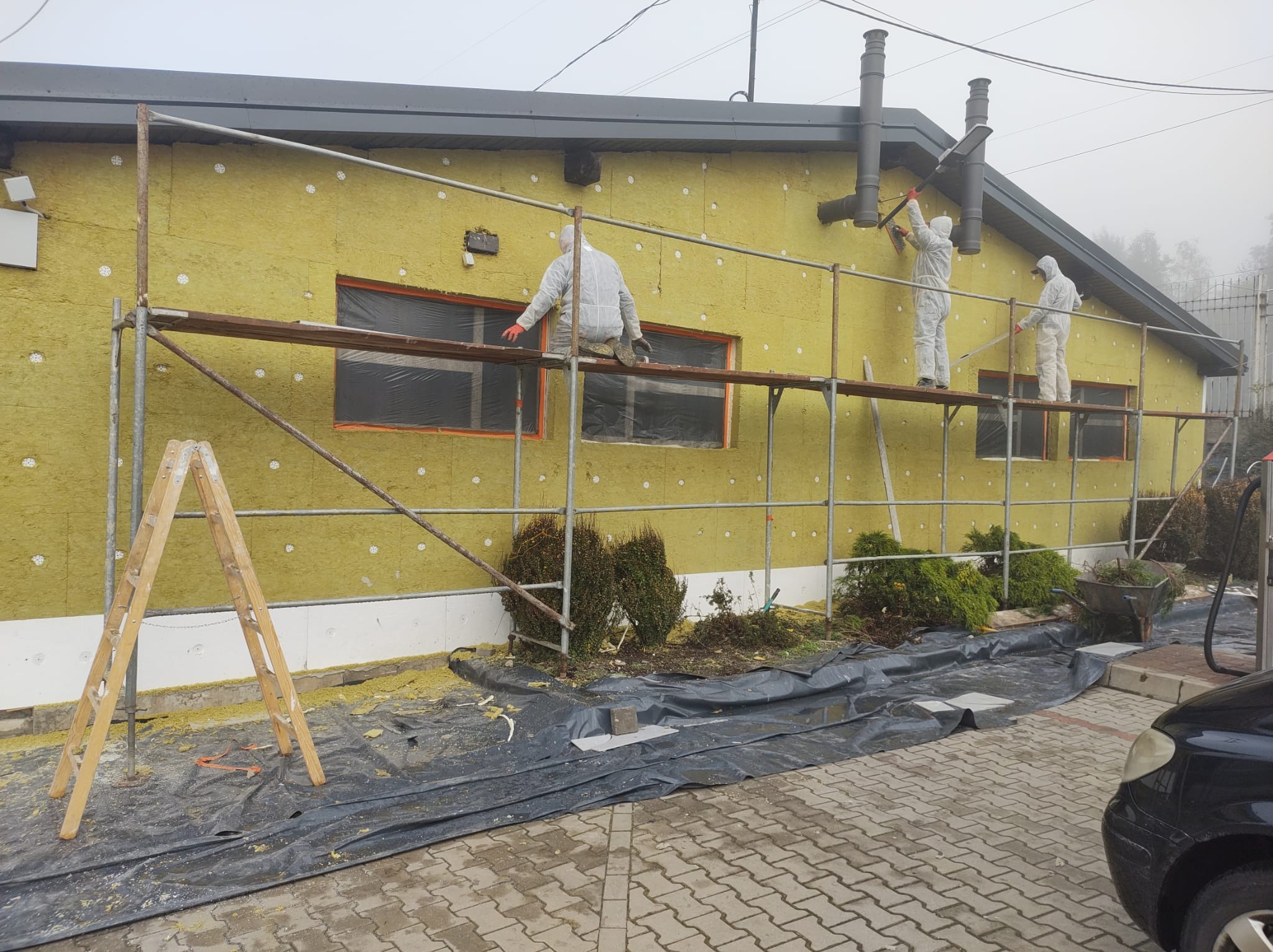 Pracownicy w kombinezonach ochronnych montują izolację termiczną na ścianie budynku, widoczne rusztowanie, zabezpieczone okna, fragment ogrodu i samochodu, pochmurny dzień.