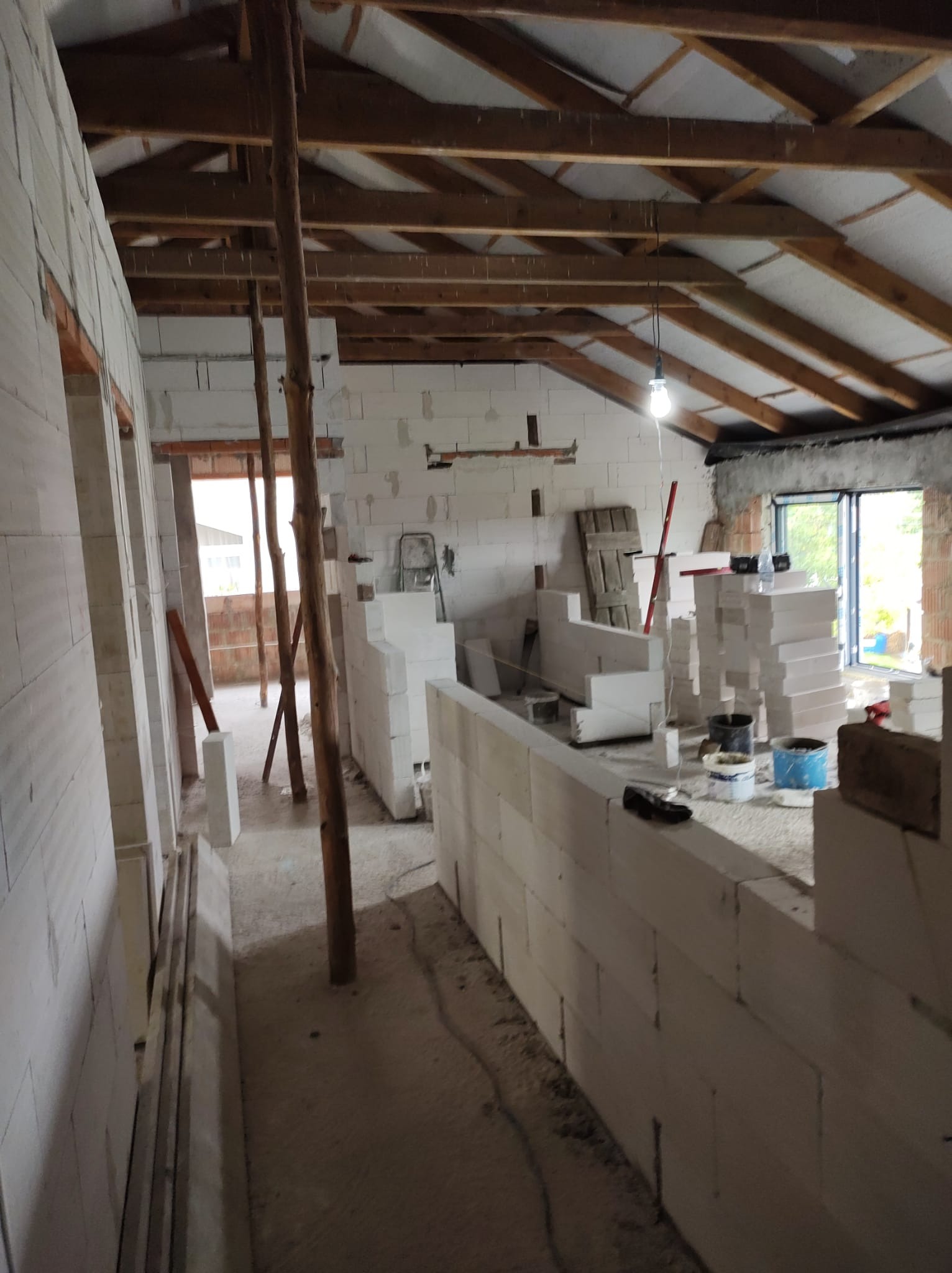 Wnętrze budynku w trakcie budowy ścian z bloczków betonu komórkowego, widoczne drewniane podpory, surowy strop z ociepleniem, okno i prowizoryczne oświetlenie.