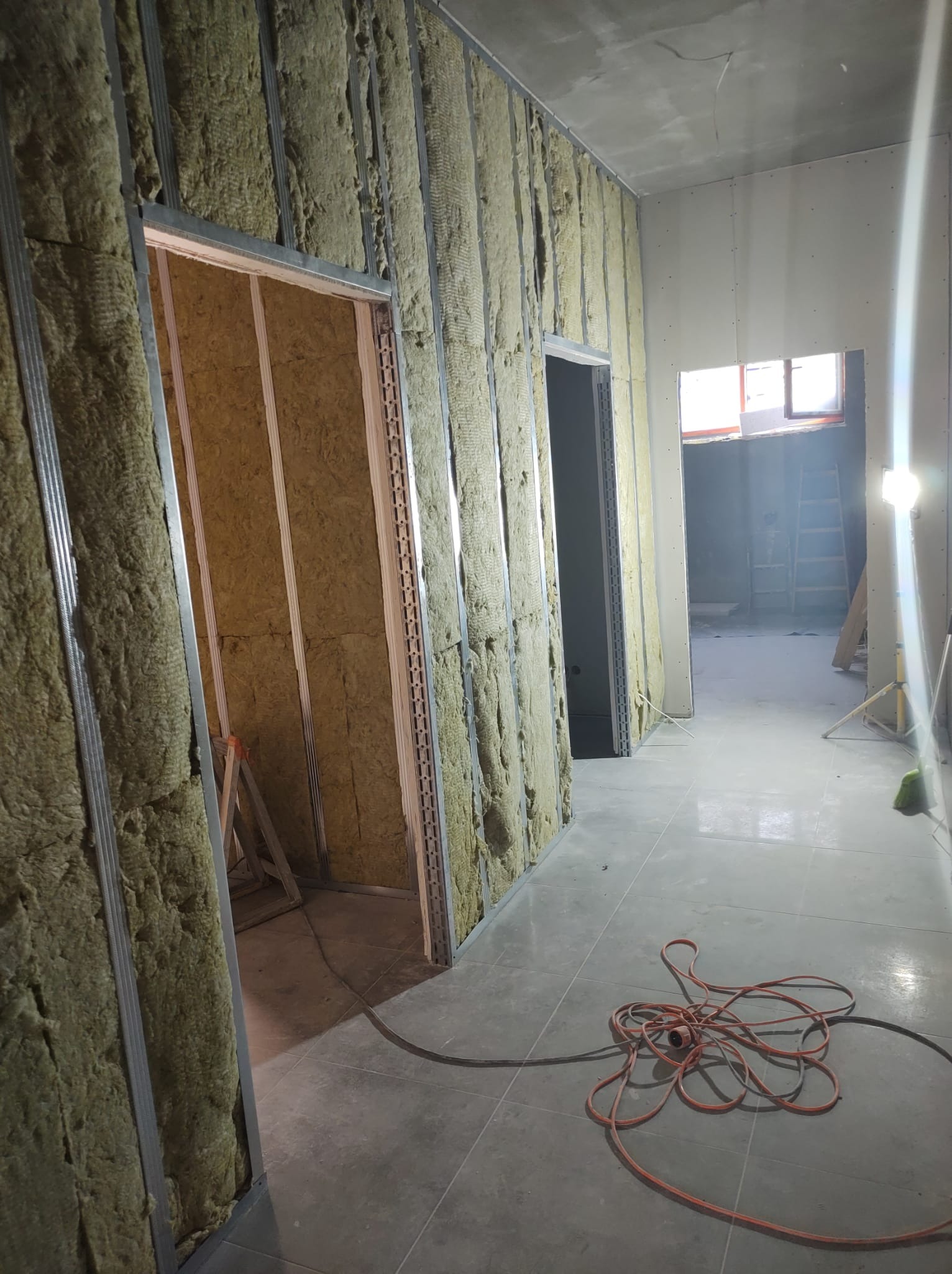 Wnętrze w trakcie ocieplania ścian wełną mineralną, widoczne metalowe profile, częściowo zamontowane płyty gipsowo-kartonowe i rozłożony kabel zasilający na podłodze.