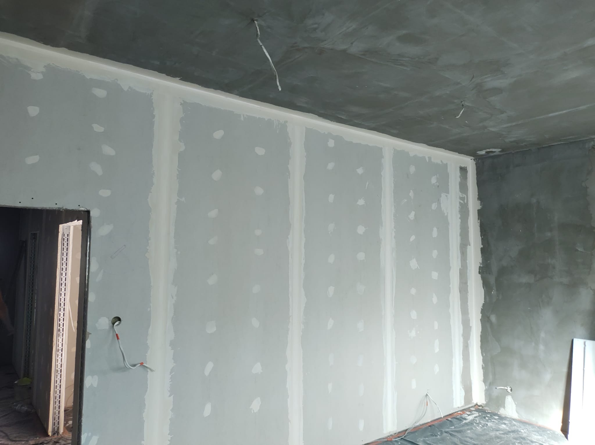 Ściana z płyt gipsowo-kartonowych w trakcie szpachlowania, widoczne łączenia płyt i otwory na kable, surowy betonowy sufit z wystającymi przewodami, przygotowanie do dalszych prac wykończeniowych.