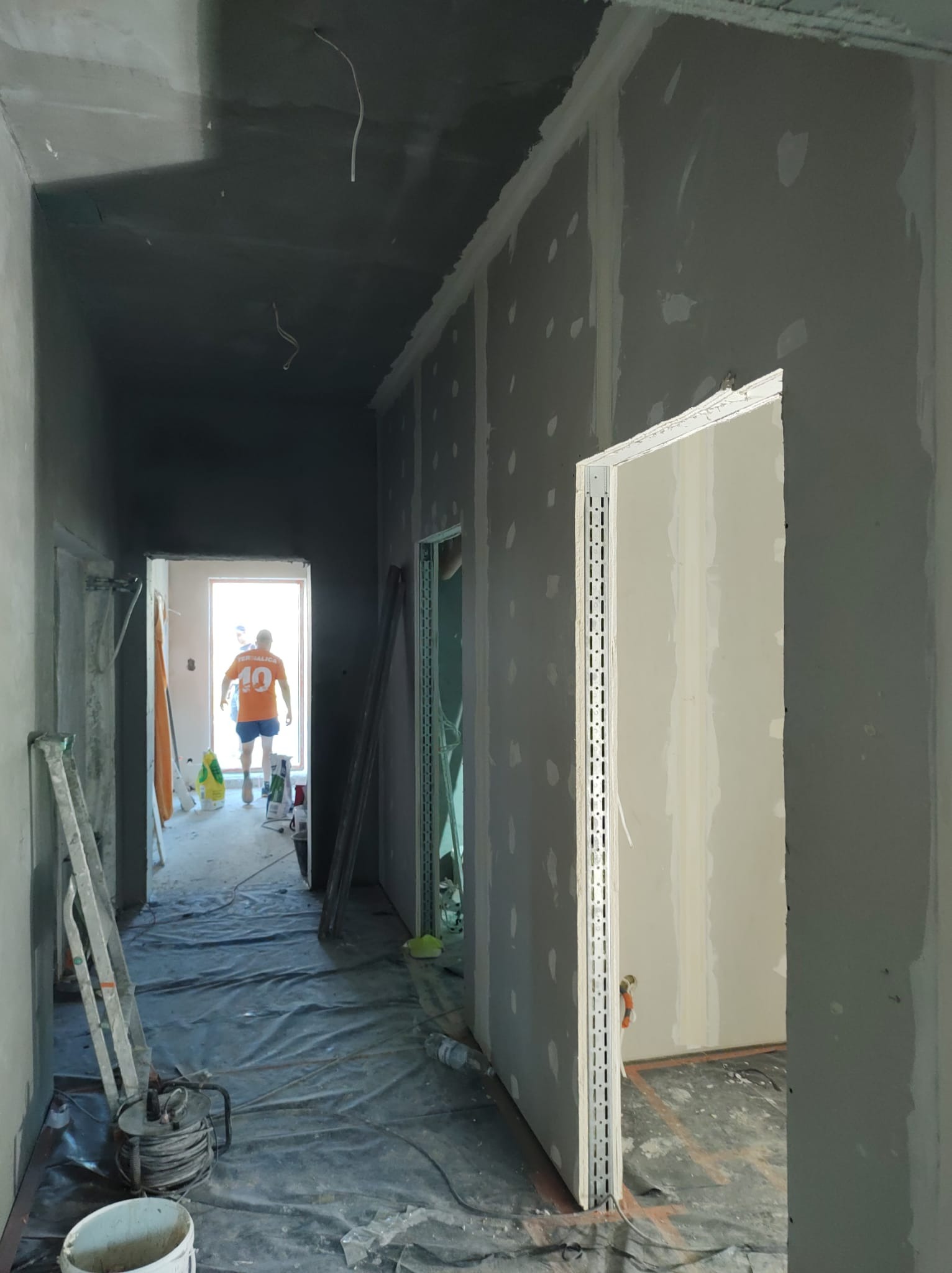 Korytarz w trakcie prac wykończeniowych: szpachlowane ściany z płyt gipsowo-kartonowych, zabezpieczona folią podłoga, widoczne kable i drabina. W tle sylwetka osoby w pomarańczowej koszulce.