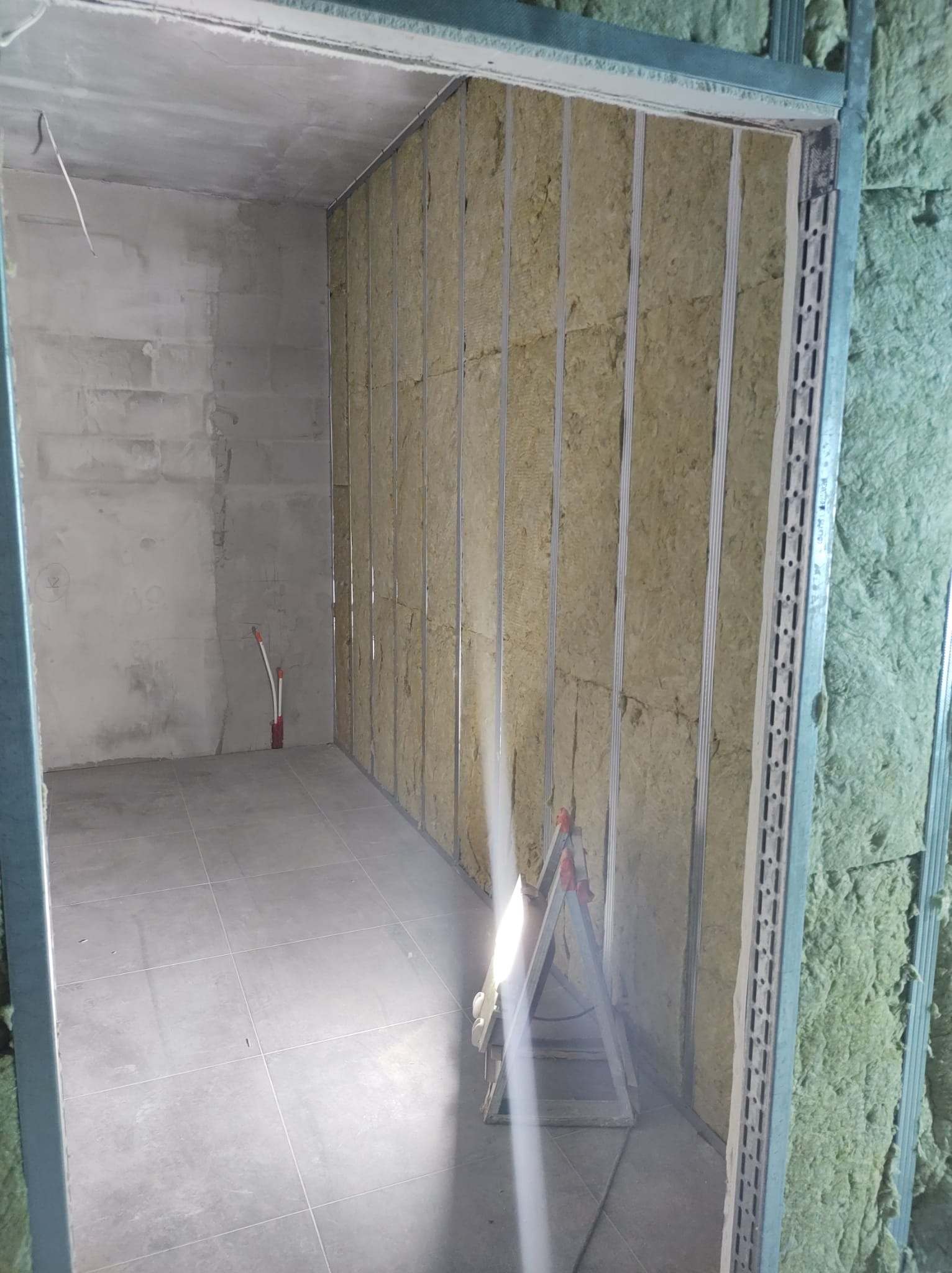 Wnętrze w trakcie ocieplania: ściana z wełną mineralną w metalowym stelażu, widoczne rury instalacji, podłoga wyłożona szarą płytką i fragment rusztowania oświetlonego lampą budowlaną.