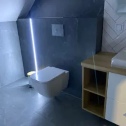 Nowoczesna łazienka z podwieszaną toaletą, szarą płytką imitującą kamień, oświetleniem LED w pionowej szczelinie i szafką umywalkową z drewnianym blatem.