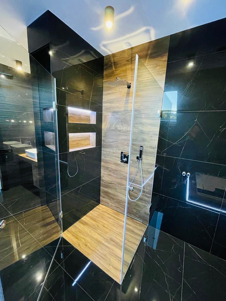 Nowoczesna kabina prysznicowa walk-in z czarnymi płytkami imitującymi marmur i drewnopodobną ścianą, podświetlona listwą LED w rogu, widoczna umywalka w tle odbijająca się w szkle.