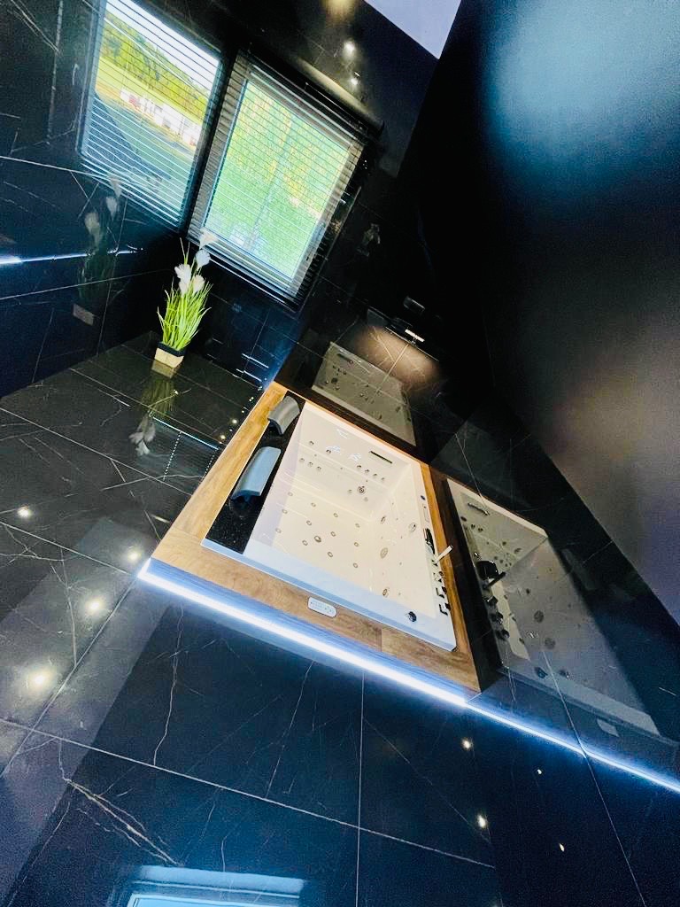 Nowoczesna łazienka z czarnymi marmurowymi płytkami, białą wanną z hydromasażem obudowaną drewnem i podświetleniem LED, widok z góry pod kątem.