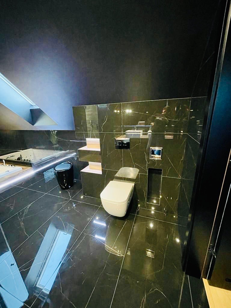 Elegancka łazienka na poddaszu z czarnymi płytkami imitującymi marmur, białą armaturą, podświetlaną wanną i czarnym koszem na śmieci.