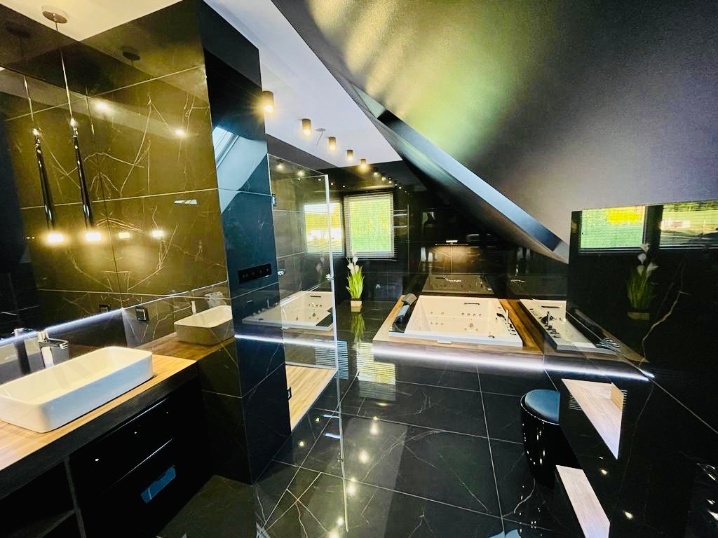 Luksusowa łazienka na poddaszu z czarnymi marmurowymi płytkami, dwiema białymi wannami jacuzzi w drewnianej obudowie, umywalką nablatową i nowoczesnym oświetleniem.