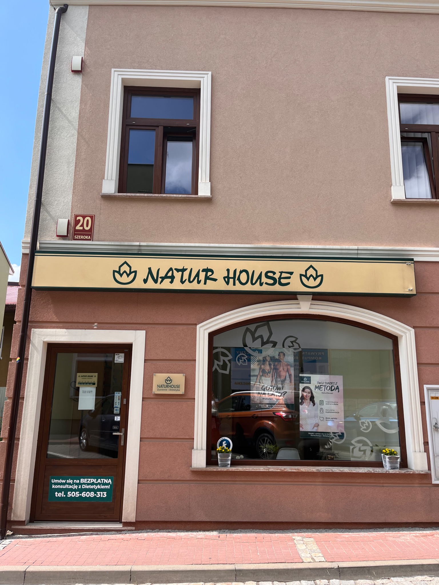 Fasada budynku z szyldem 'Natur House' z numerem 20 przy ulicy Szerokiej, informacja o bezpłatnej konsultacji z dietetykiem na drzwiach wejściowych, okno wystawowe z plakatami reklamowymi...