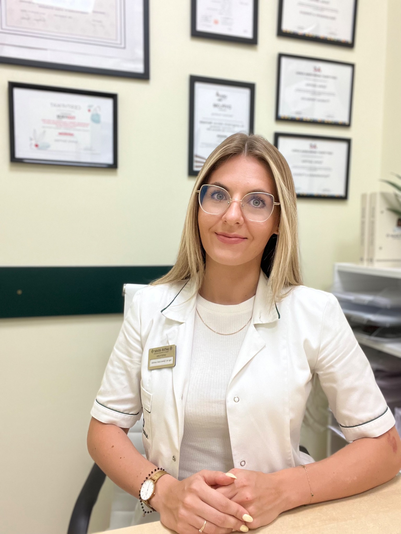 Kobieta w białym fartuchu lekarskim i okularach siedzi przy biurku w gabinecie, za nią na ścianie wiszą oprawione dyplomy.