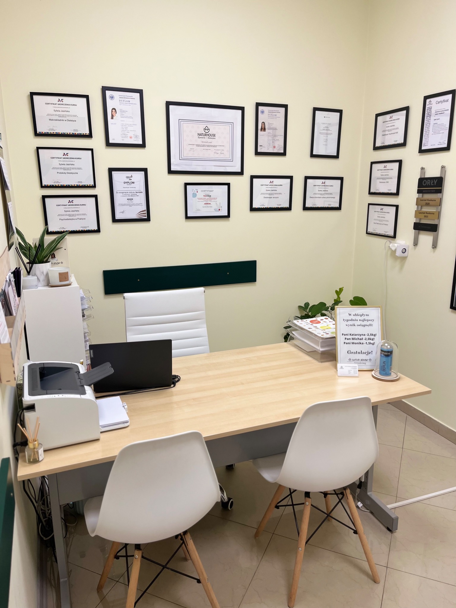 Gabinet z biurkiem, dwoma krzesłami i ścianą pełną certyfikatów oraz dyplomów, w tym wyróżnienie 'Orły Medycyny', informacja o najlepszych wynikach pacjentów w odchudzaniu w ostatnim tygodniu.