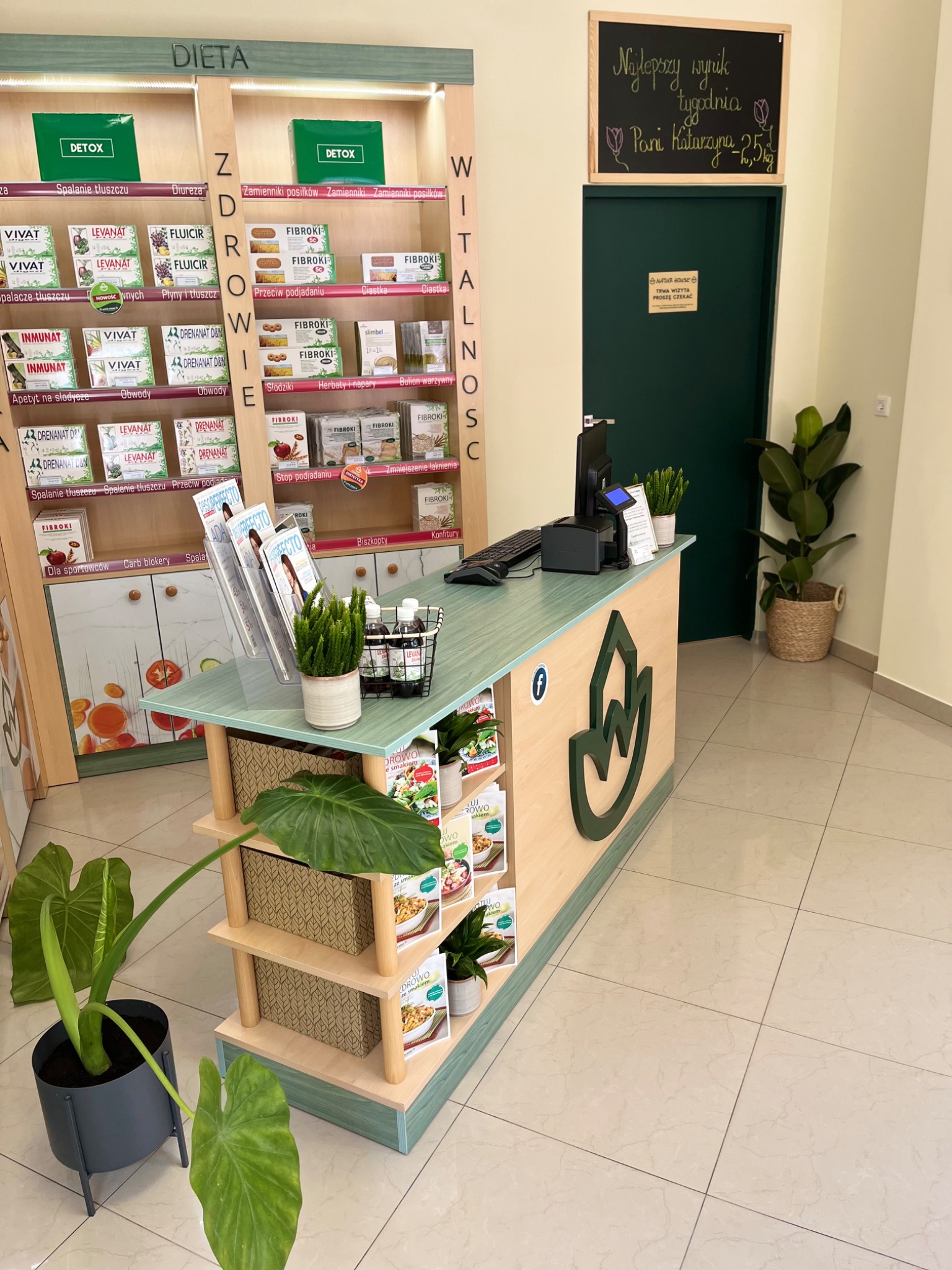 Wnętrze gabinetu z regałem pełnym suplementów diety, stanowiskiem obsługi klienta z zielonym logo i tablicą z napisem 'Najlepszy wynik tygodnia'.