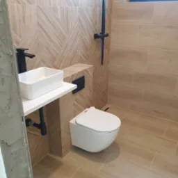 Nowoczesna łazienka z beżowymi płytkami imitującymi drewno, białą umywalką nablatową na białym blacie, czarną armaturą i wiszącą toaletą.