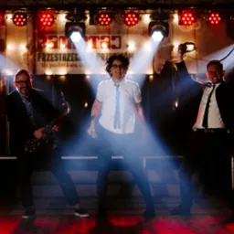 Zespół muzyczny w trakcie występu na scenie: saksofonista, wokalistka z mikrofonem i mężczyzna z słuchawkami, oświetlenie sceniczne i czerwone światła.