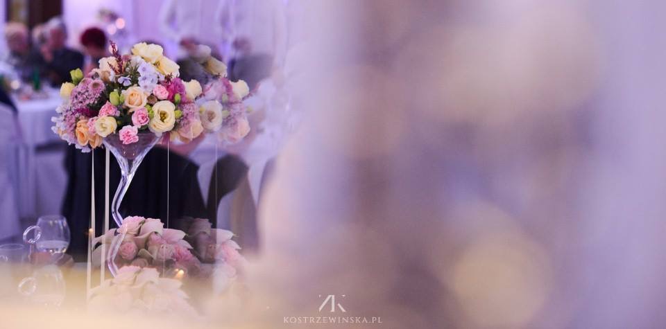 Kompozycja kwiatowa w szklanym wazonie martini na stole weselnym, z delikatnym oświetleniem i rozmytym tłem gości.