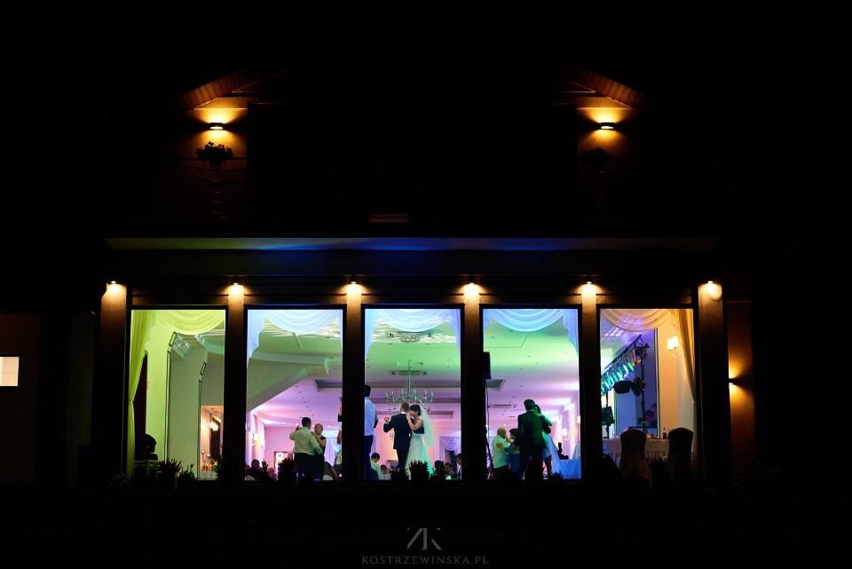 Nocne ujęcie sali balowej z zewnątrz, widoczne tańcząca para młoda, dekoracje świetlne w odcieniach zieleni, fioletu i błękitu, oraz białe draperie okienne.