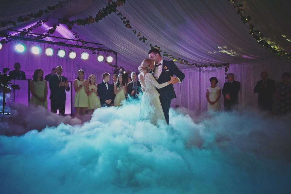 Para młoda tańczy pierwszy taniec w chmurze dymu na tle oświetlonej sceny z gośćmi weselnymi.